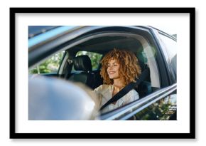 Smiling woman driving car fastened with seatbelt, curly hair, casual clothing, daylight, safe urban travel, joyful expression, natural light.