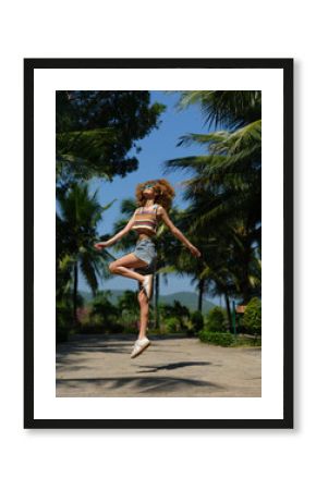 Smiling woman jumps outdoors wearing colorful top and denim shorts under palm trees on sunny summer day with blue sky background.