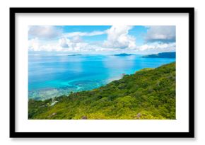 Aerial view of Praslin Island, Seychelles, featuring lush green hills, tropical coastline, and calm turquoise waters under a partly cloudy sky. 