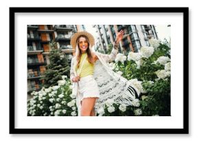 Young woman in sunhat and white crochet shawl joyful in a sunny city garden among white hydrangeas