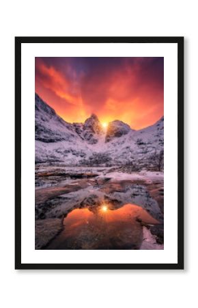 Vibrant orange and red sunrise illuminating snow-covered peaks, reflecting on the icy surface of frozen lake in a tranquil winter mountain landscape. Snowy mountains and frozen fjord. Lofoten, Norway