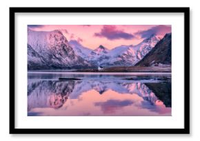 Beautiful pink sunrise illuminating snowy mountains and houses reflecting in the calm water of a fjord in Lofoten islands, Norway. Winter landscape with sea, pink sky with clouds, rorbu, rocks in snow