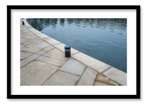 Small cylindrical outdoor light fixture on granite pavement near water surface. Landscape lighting design, waterfront infrastructure, and architectural illumination systems. Real photo.