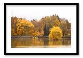 This tranquil pond in Kudowa Zdroj features vibrant autumn colors with shades of orange and yellow. Visitors stroll along the water's edge, enjoying the peaceful fall atmosphere.