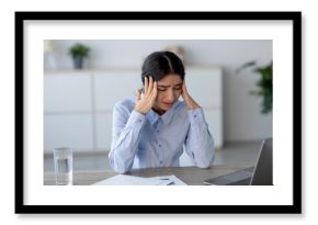 A young Indian woman sits at her workplace, visibly upset and holding her head. She appears to be suffering from a headache while surrounded by papers and a laptop, indicating work-related stress.