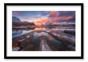 Arctic landscape with snowy mountains reflecting on calm lake with stones and colorful sky with clouds at sunrise. Winter in Lofoten islands, Norway. Sea, rocks in snow, pink sky with clouds at dusk