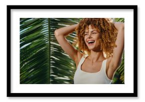 Smiling woman with curly hair enjoys tropical sunlight wearing white tank top under palm leaves, expressing happiness and natural beauty in vibrant outdoor setting.