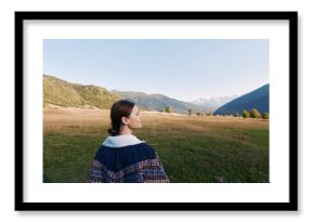 Woman stands in meadow valley looking at mountains and nature landscape outdoors, travel portrait in sweater with serene scenery, grass, sky and panoramic alpine view.