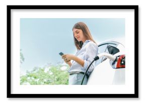 Young woman using smartphone online banking application to pay for electric car battery charging from EV charging station during vacation road trip at national park or summer forest. Panorama Exalt