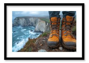 A pair of sturdy yellow hiking boots stands on the edge of a coastal cliff, overlooking turbulent ocean waves beneath a cloudy sky. The landscape evokes adventure.