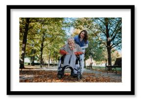 Disabled elderly woman in wheelchair on walk in the park during warm autumn day. Young nurse pushing wheelchair, talking with senior woman.