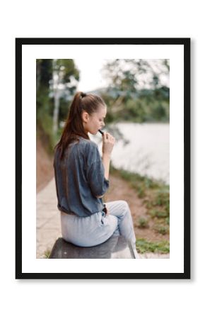 Thoughtful young woman sitting by the river, enjoying her vape while contemplating life Peaceful outdoor setting with nature and calm waters