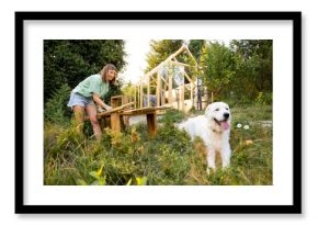 Female carpenter standing by a workbench and her large white dog in front of a partially built wooden greenhouse in her backyard. Engaged in a DIY project, surrounded by nature