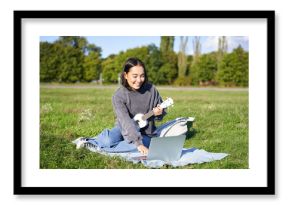 Happy asian girl plays ukulele outdoors, teaches music online with her laptop, sitting in park, playing instrument