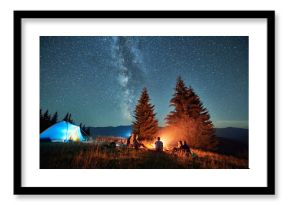 Night camping in mountains under starry sky. Group of people tourists having a rest near campsite, burning campfire and illuminated tent. Concept of tourism, hiking and adventure.