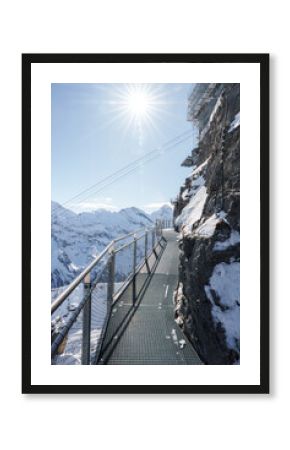 Metal walkway on rocky cliff at Murren ski resort, Switzerland. Snow covered mountains in distance, clear blue sky, and safe pathway for exploring Alps.