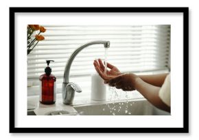 Hands under running water at kitchen sink with soap dispenser and yellow flowers in background, focusing on hygiene and cleanliness