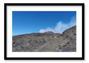 Rocky volcanic terrain with scattered lava stones and slopes surrounding a smoking crater on Mount Etna beneath a vivid blue sky in Sicily, Italy
