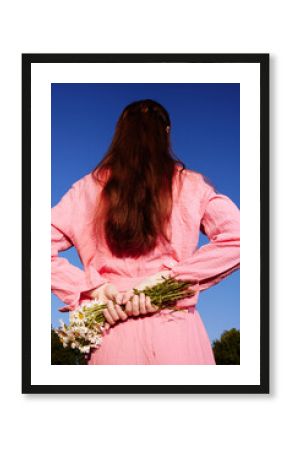 A young woman holding flowers behind her back, wearing a pink outfit, with long brown hair, standing against a bright blue sky, evoking feelings of innocence and joy