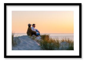A couple sits on a sandy dune, surrounded by tall grass, admiring the vibrant sunset over the calm waters of the beach in Denmark. The warm hues create a romantic atmosphere. Vejers Strand beach