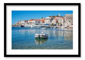 A picturesque coastal town of Primosten in Croatia features clear turquoise water, moored yachts, stone houses with red rooftops, and a peaceful Mediterranean atmosphere under a bright blue sky.