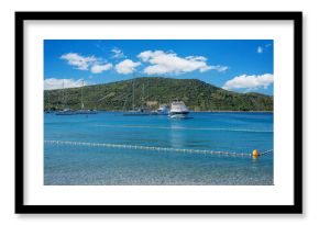 A tranquil coastal scene featuring clear turquoise waters, anchored yachts, and a green hillside under a bright blue sky in Primosten, Croatia.