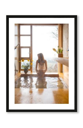 A woman enjoys a serene bath in a wooden tub by a panoramic window. Surrounded by flowers and soft daylight, the moment captures peaceful self-care and quiet luxury