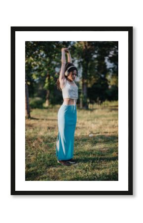 A young woman enjoys a sunny day in the park as she stretches her arms overhead, listening to music. She appears relaxed and focused, embracing the outdoors.