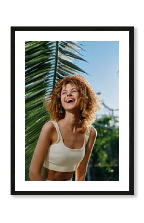 smiling woman with curly hair wearing white crop top outdoors under sunny sky by tropical palm leaves, joyful and relaxed, vibrant summer atmosphere