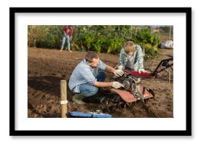 Male farmer shows his son how to fix garden cultivator. Farmer and a guy are repairing a machine to cultivate the soil with tools. Breakdown and repair of garden equipment on the farm