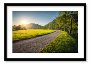 Sun setting behind mountains illuminating gravel road winding through green grass field next to a river with trees and forest in the background during a beautiful spring evening in Slovenia. Sunset