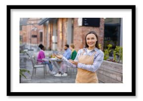 Smiling waitress welcomes customers to an outdoor restaurant, gesturing towards the seating area with other patrons.