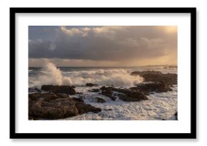 Rough seas crash against rocky shores along the Whale Coast, Hermanus, in the scenic Overberg region of Western Cape, South Africa, under dramatic cloudy skies and vibrant coastal vegetation.