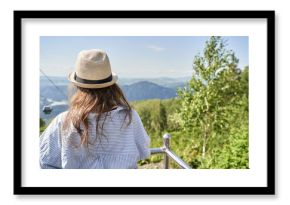 Female in straw hat enjoying scenic mountain view from hilltop balcony on a sunny day.