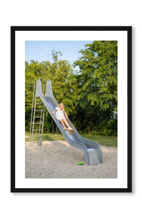 Preschooler having fun sliding down a metal slide on a playground during a bright summer day, surrounded by green trees