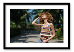 young woman with curly hair wearing colorful crochet top and denim shorts standing outdoors, enjoying sunny weather, smiling with eyes closed, tropical background