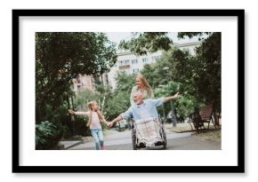 Joyful family outing in the park as grandpa in wheelchair shares a smile with granddaughter and daughter guiding them along a sunny nature path