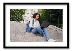 Young woman student sits on campus stairs with laptop smiling outdoors in urban park setting
