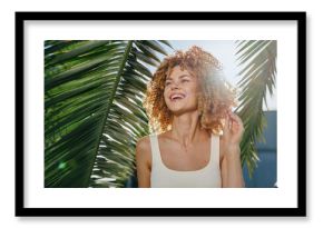 smiling woman with curly hair enjoying sunny tropical outdoors, wearing white tank top framed by green palm leaves and bright sunshine