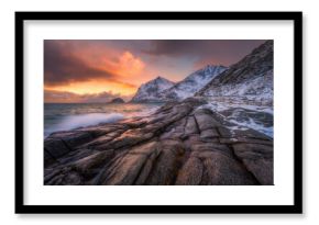 Dramatic winter seascape on the Lofoten Islands in Norway with waves crashing over coastal rocks and snow-covered mountains at sunset. Arctic landscape with sea, stones, blurred water, colorful sky