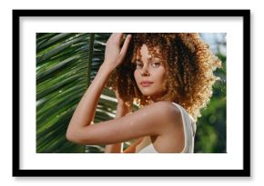 Curly-haired woman posing outdoors in white top near tropical palm leaves with natural sunlight and green blurred background under warm summer conditions.
