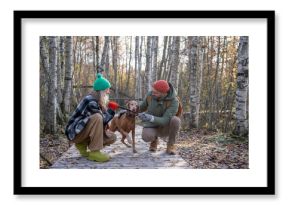 Smiling couple sitting on haunches on wooden walkway playing with magyar vizsla, training hound dog. Clever puppy holding stick in teeth, refusing to drop. Spending pastime in nature together with pet