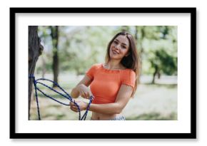 A young woman smiling as she sets up a hammock in a green park on a sunny day. The atmosphere is cheerful and relaxed.