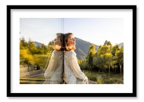 A woman sits quietly by a window, gazing at the surrounding mountains and forest. The serene landscape and reflection create a perfect moment of solitude and connection with nature