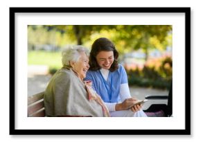 Portrait of nurse and elderly woman on walk in the park during warm autumn day. Young caregiver looking through photos of senior patient's family on a phone.