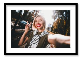 Photo of pretty young blonde woman take selfie photo show v-sign wear khaki clothes tied shoulders sweater walk outside urban city park