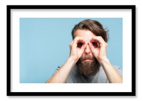 funny ludicrous joyful comic playful man pretending to look through binoculars made of hands. portrait of a young bearded guy on blue background. emotion facial expression concept