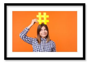 Popularity in social media. Portrait of funny brunette woman wearing checkered casual shirt holding large big yellow hashtag sign on head as crown. indoor studio shot isolated on orange background