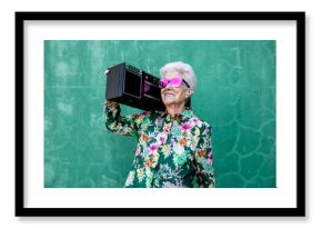 Cool elderly female in colorful trendy blouse and sunglasses carrying record player and enjoying music against green wall