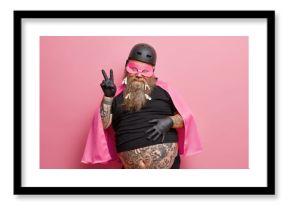 Portrait of funny bearded superhero makes peace gesture has big tattooed belly wears mask helmet and cape isolated over pink background. European adult man shows victory sign being on costume party